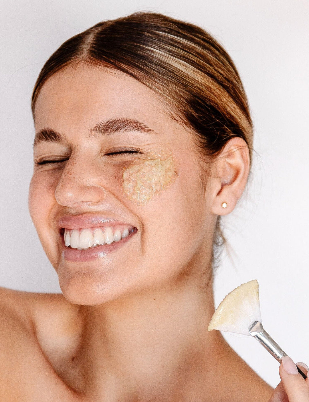 A smiling woman with a creamy exfoliant on her cheek holds a flat brush beside her face, demonstrating Biologi Bx Reveal Exfoliant.