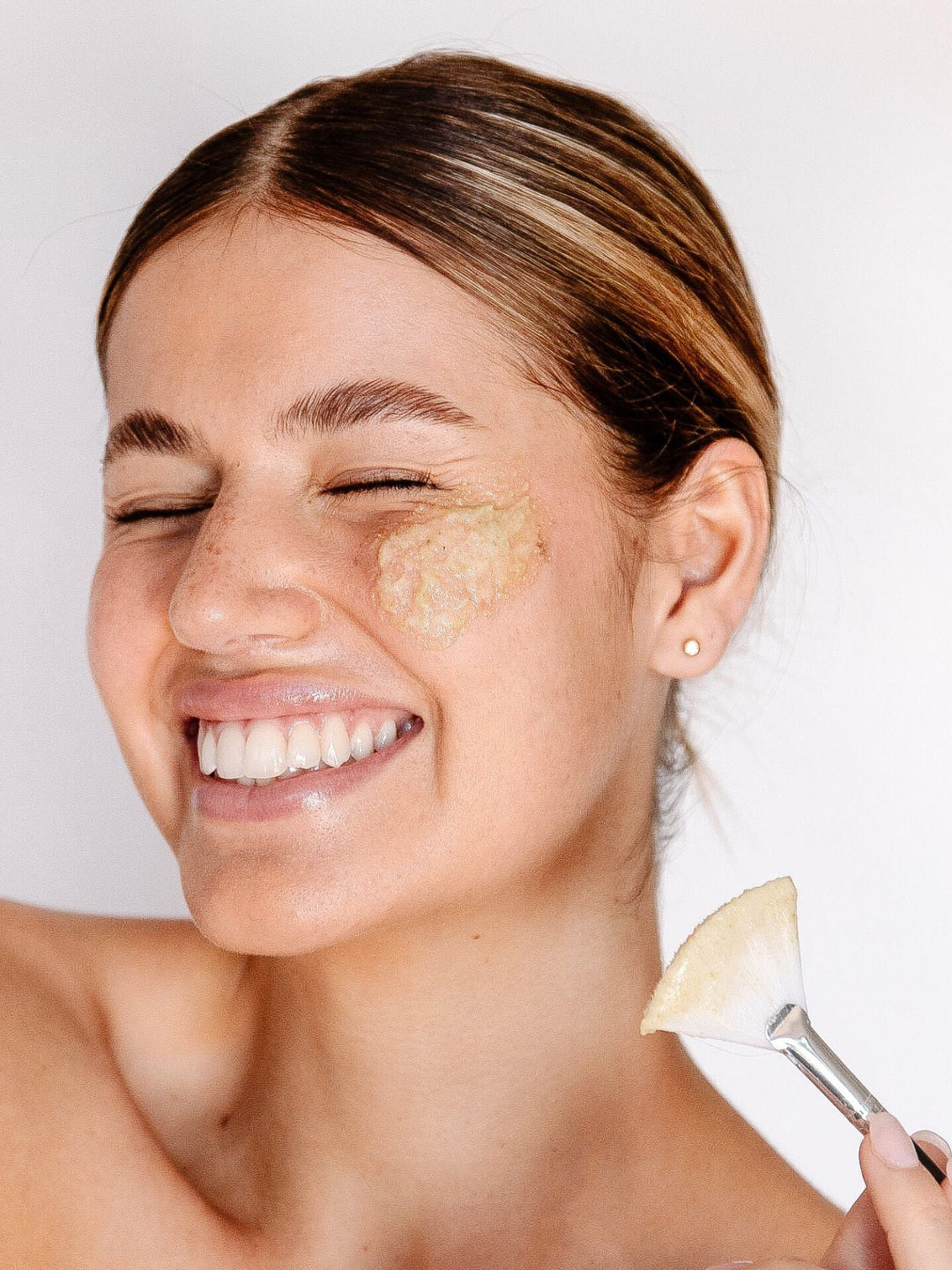 A smiling woman with a creamy exfoliant on her cheek holds a flat brush beside her face, demonstrating Biologi Bx Reveal Exfoliant.