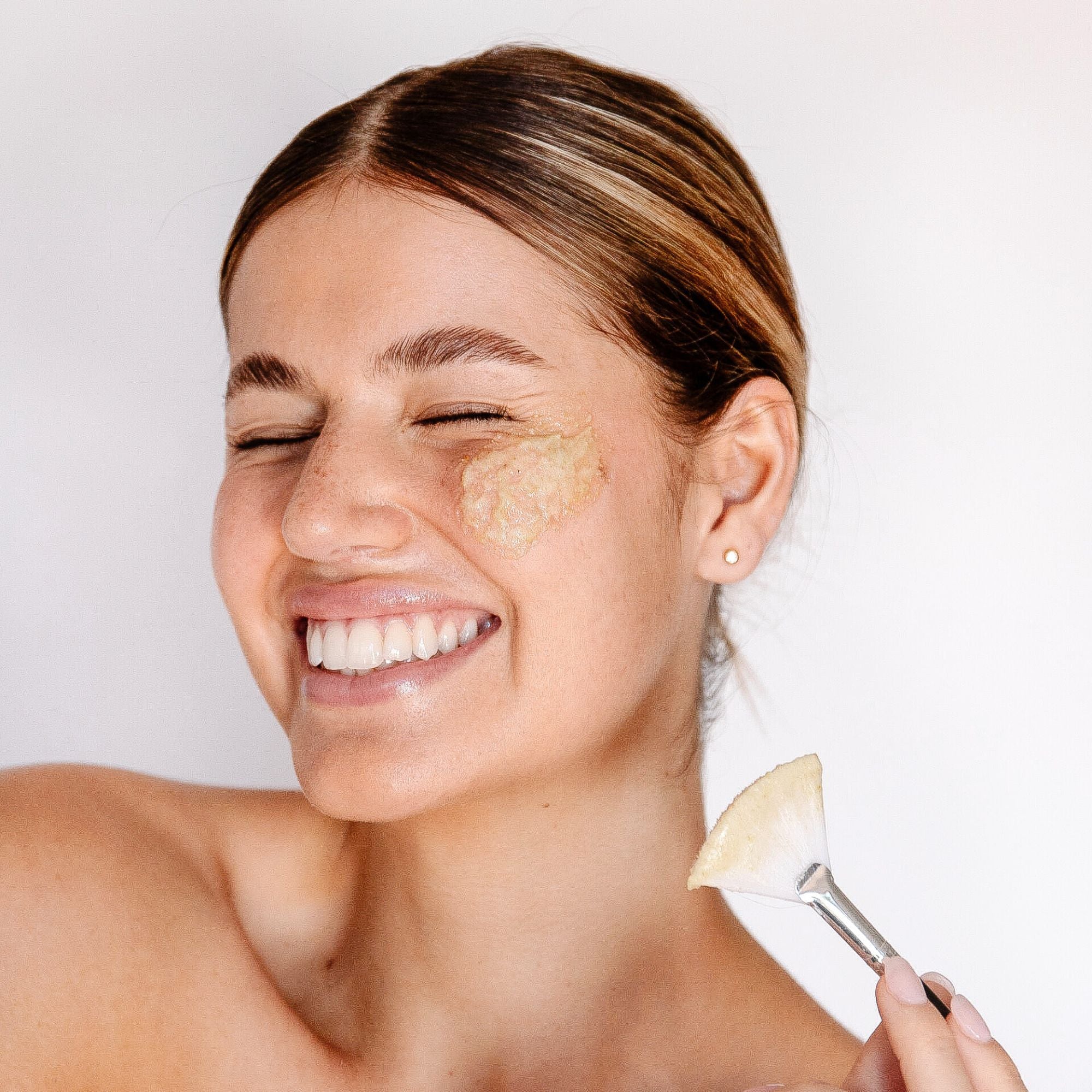 A smiling woman with a creamy exfoliant on her cheek holds a flat brush beside her face, demonstrating Biologi Bx Reveal Exfoliant.