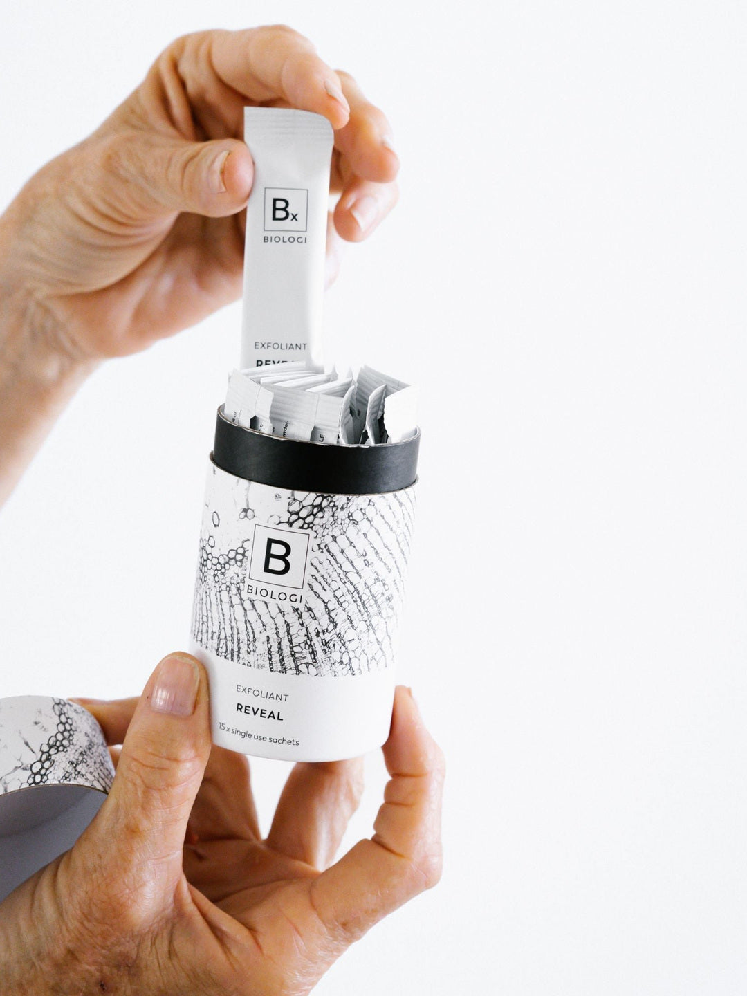 Hands lift an individual Biologi Bx Reveal Exfoliant sachet from the open cylindrical carton, showing the sachets inside and the removed lid.