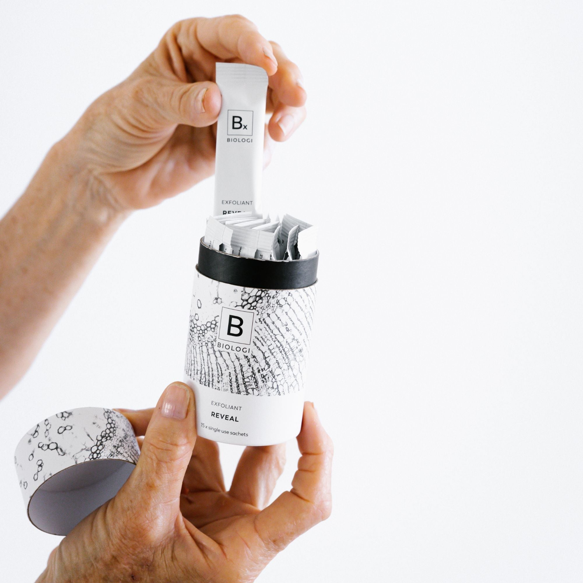 Hands lift an individual Biologi Bx Reveal Exfoliant sachet from the open cylindrical carton, showing the sachets inside and the removed lid.