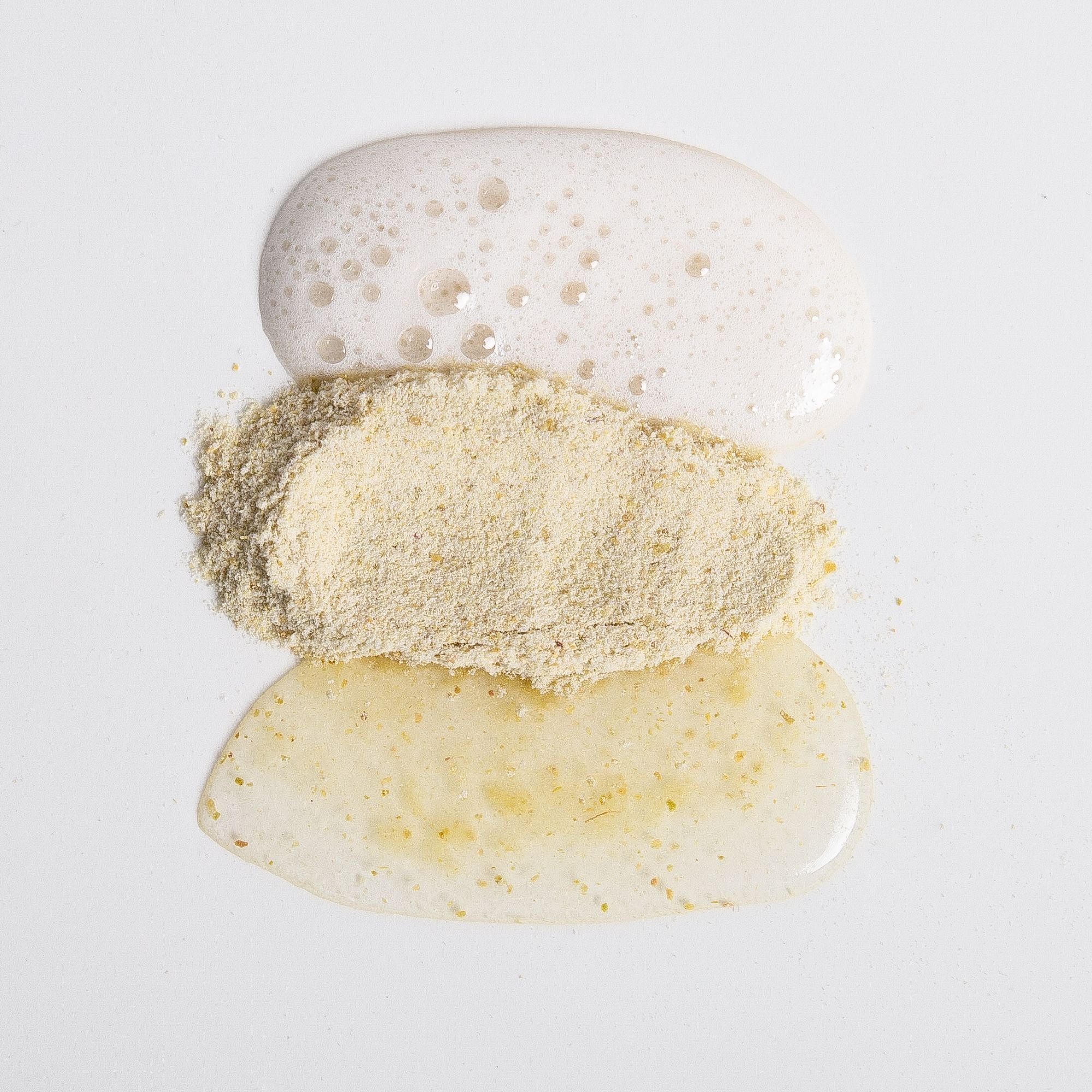 Biologi Bx Reveal Exfoliant powder between white cleanser foam and a golden mixed cream on a pale surface, showing the textures before and after mixing.