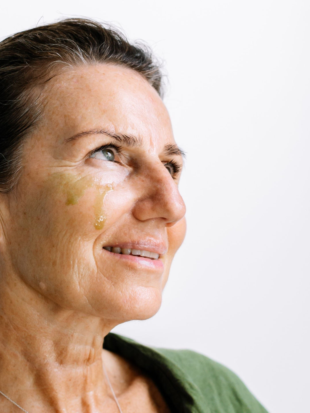 A woman looks to the side with golden Biologi By Blackberry Seed Oil visible on her cheek, showing the facial oil on skin.