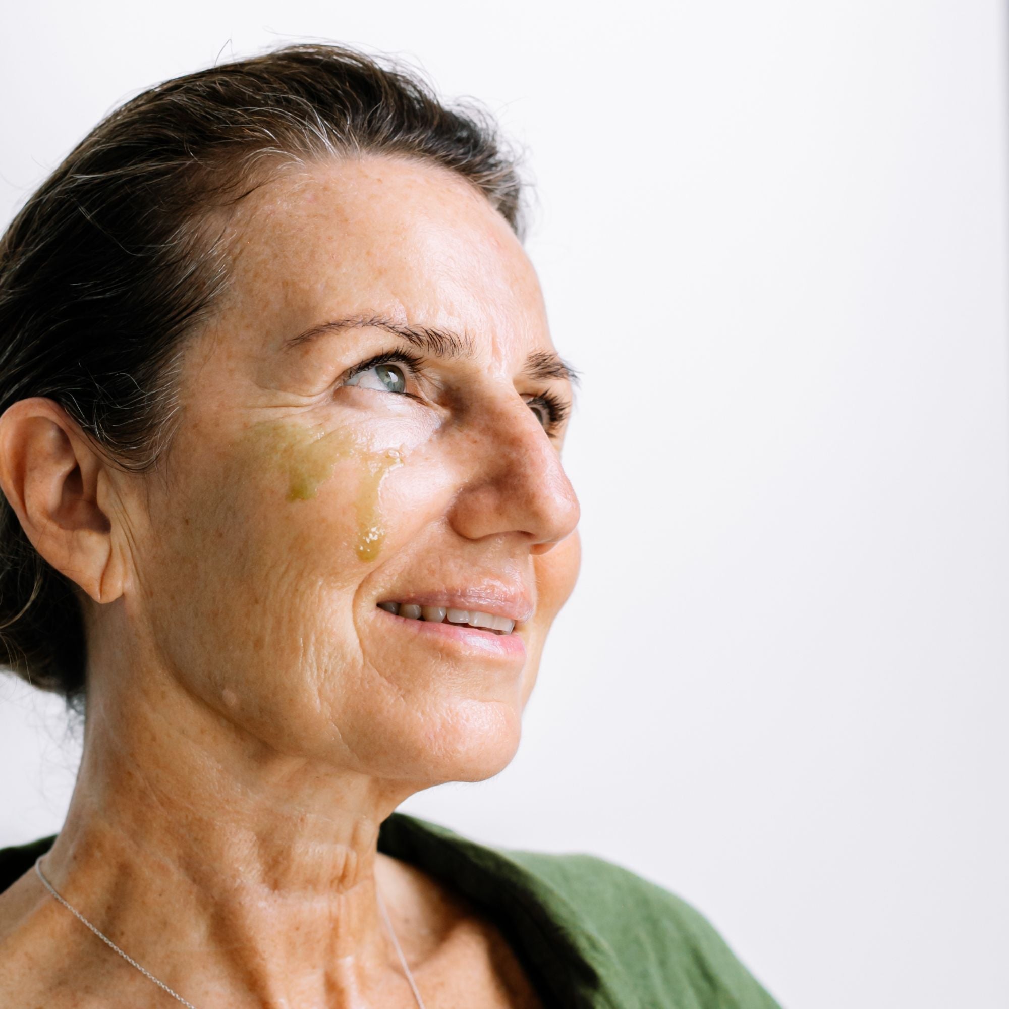 A woman looks to the side with golden Biologi By Blackberry Seed Oil visible on her cheek, showing the facial oil on skin.