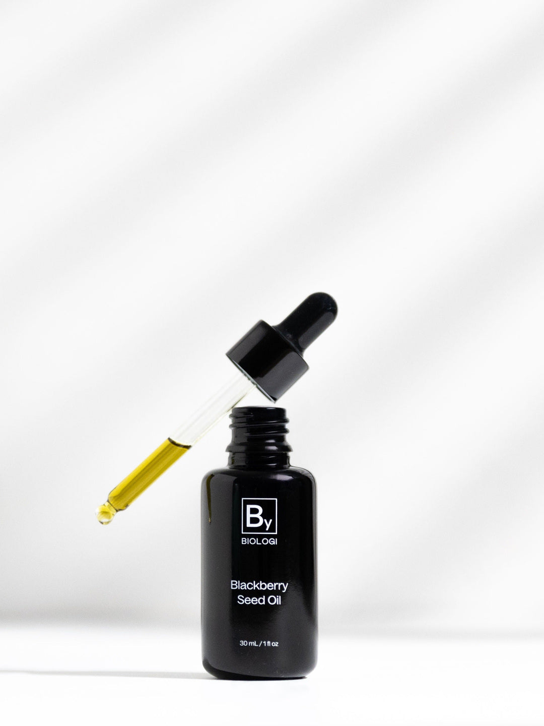 An open black Biologi By Blackberry Seed Oil bottle on white beneath a glass dropper filled with golden oil, with diagonal light and shadow behind it.