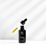 An open black Biologi By Blackberry Seed Oil bottle on white beneath a glass dropper filled with golden oil, with diagonal light and shadow behind it.