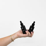 A hand holds Biologi By Blackberry Seed Oil and Br Rosehip Oil together, displaying both black dropper bottles against a pale backdrop.