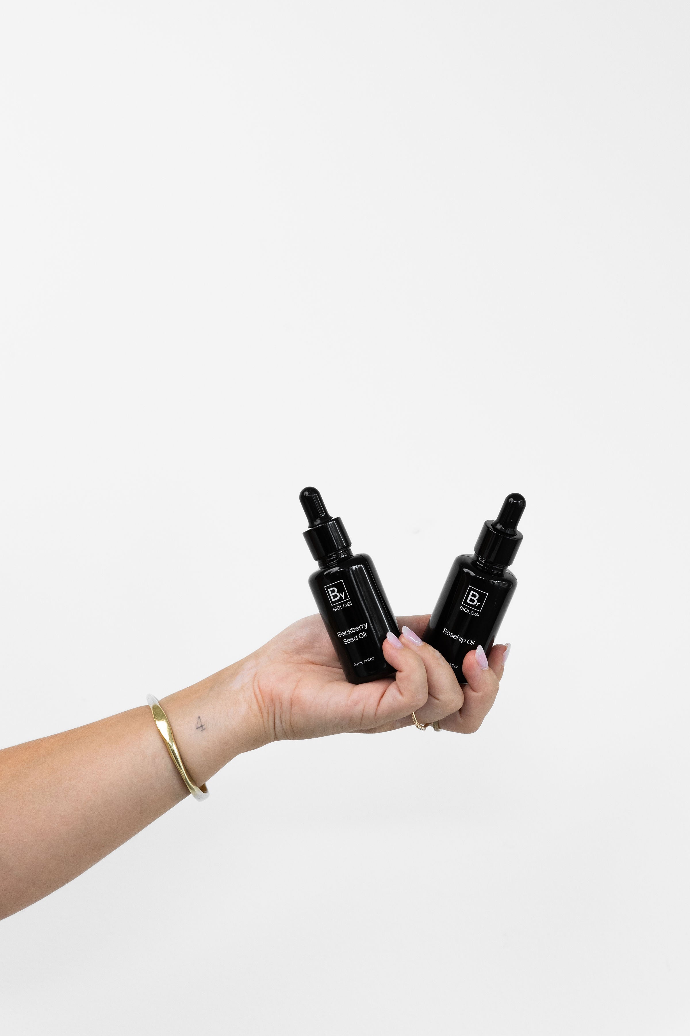 A hand holds Biologi By Blackberry Seed Oil and Br Rosehip Oil together, displaying both black dropper bottles against a pale backdrop.