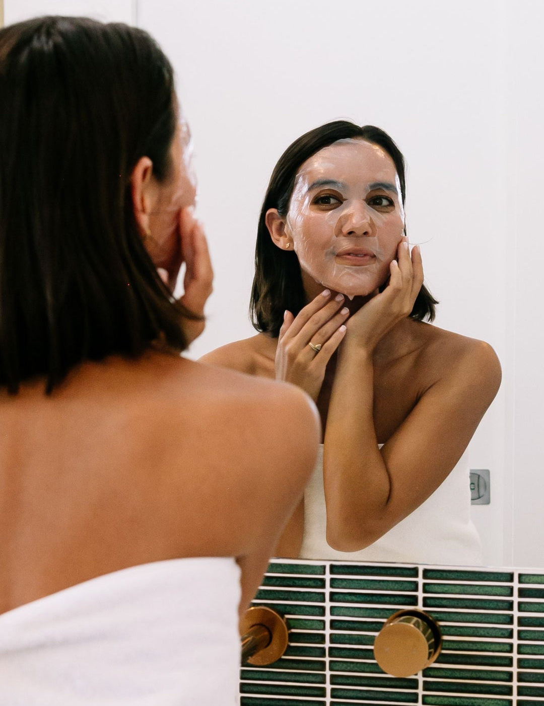 A woman wearing Biologi Hydra Balance Bio Cellulose Sheet Mask smooths it against her face while looking into a bathroom mirror.