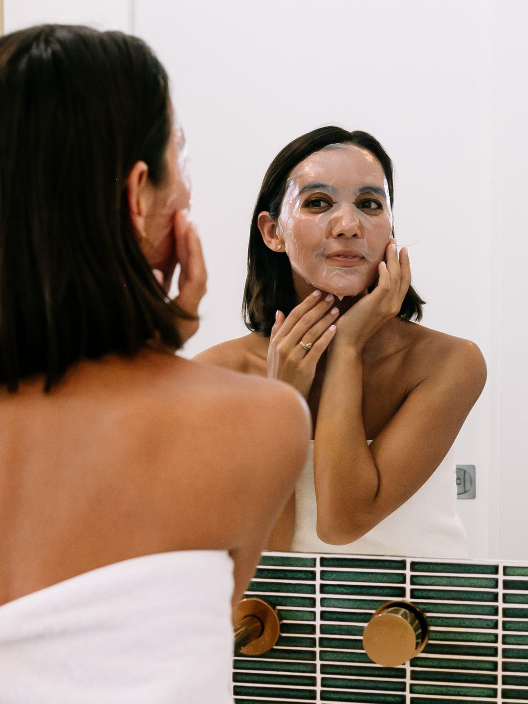A woman wearing Biologi Hydra Balance Bio Cellulose Sheet Mask smooths it against her face while looking into a bathroom mirror.