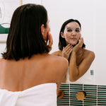 A woman wearing Biologi Hydra Balance Bio Cellulose Sheet Mask smooths it against her face while looking into a bathroom mirror.