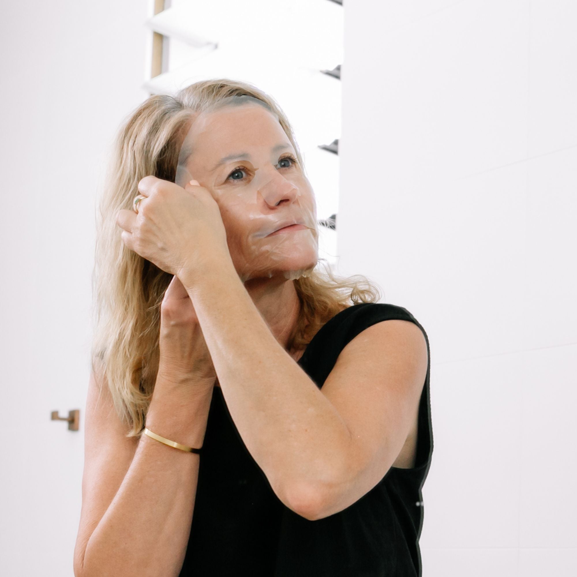 A woman in a black sleeveless top adjusts Biologi Hydra Balance Bio Cellulose Sheet Mask near her temple in a bright bathroom.