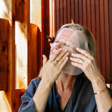 A woman presses a translucent Biologi Hydra Boost Bio Cellulose Sheet Mask onto her face with both hands, seated beside a warm timber wall.