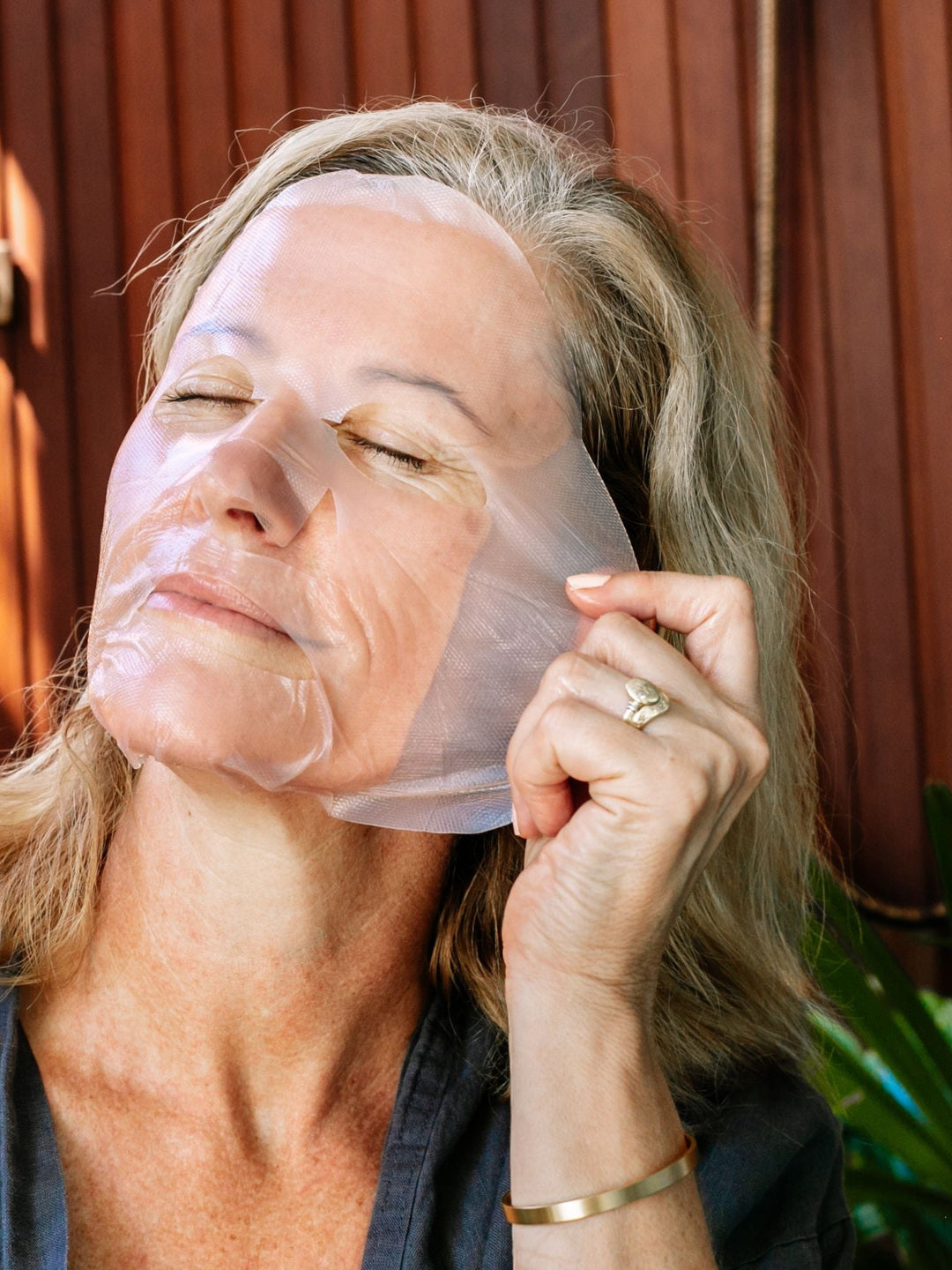 A woman wearing the Biologi Hydra Boost Bio Cellulose Sheet Mask adjusts its edge beside her ear, with her eyes closed against a timber backdrop.