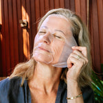 A woman wearing the Biologi Hydra Boost Bio Cellulose Sheet Mask adjusts its edge beside her ear, with her eyes closed against a timber backdrop.