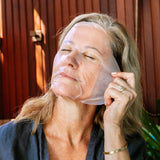 A woman wearing the Biologi Hydra Boost Bio Cellulose Sheet Mask adjusts its edge beside her ear, with her eyes closed against a timber backdrop.