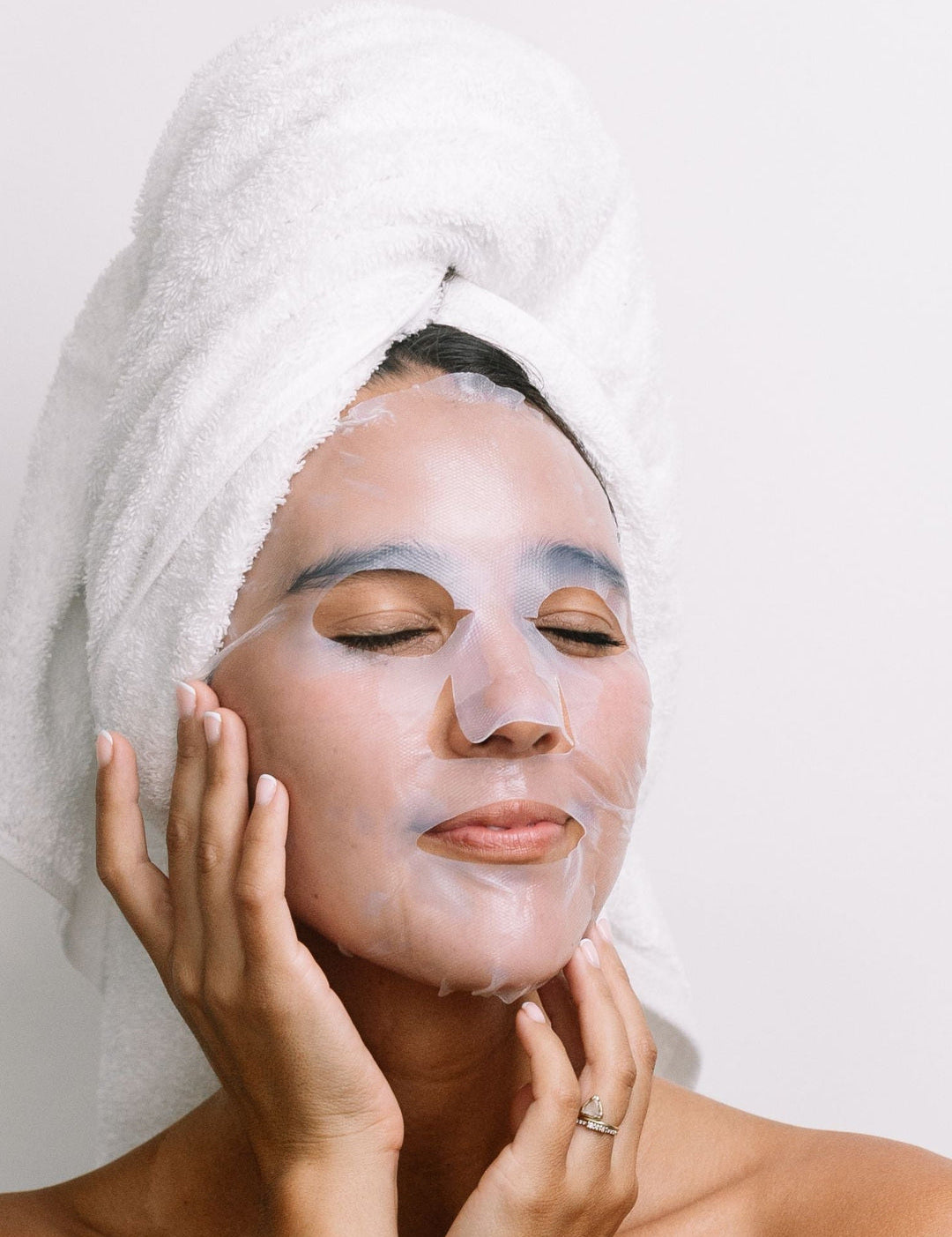 A woman with a white towel around her hair wears the translucent Biologi Hydra Calm Bio Cellulose Sheet Mask and gently rests her hands against her face.