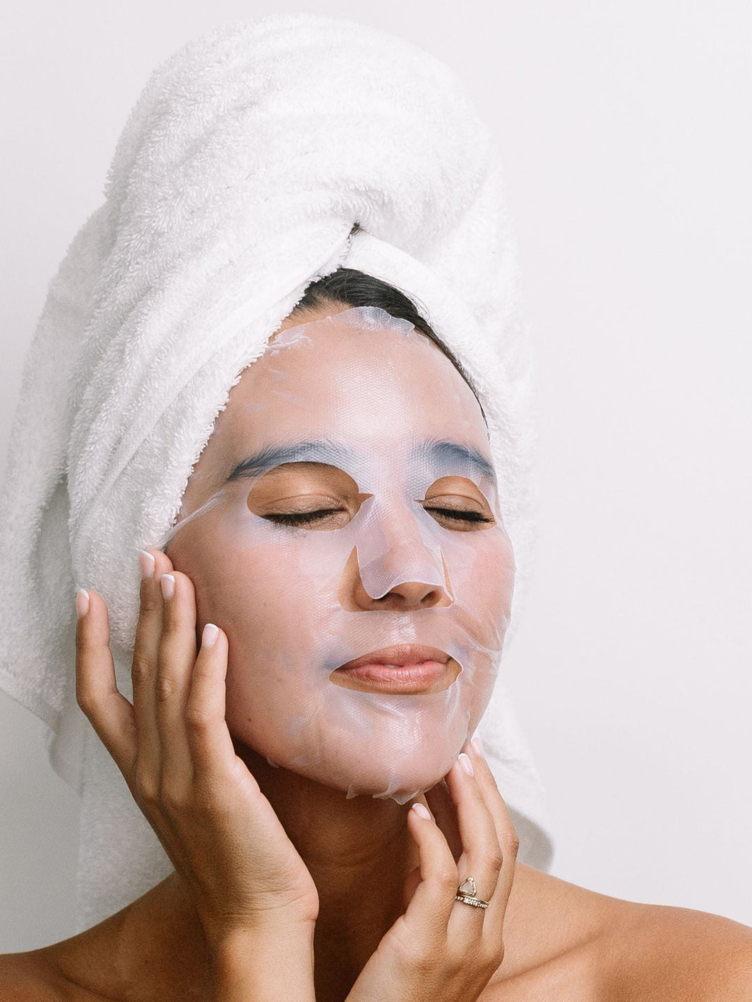 A woman with a white towel around her hair wears the translucent Biologi Hydra Calm Bio Cellulose Sheet Mask and gently rests her hands against her face.