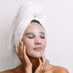 A woman with a white towel around her hair wears the translucent Biologi Hydra Calm Bio Cellulose Sheet Mask and gently rests her hands against her face.
