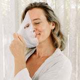 A woman in a white robe lifts a translucent Biologi Hydra Calm Bio Cellulose Sheet Mask towards her face beside softly lit curtains.