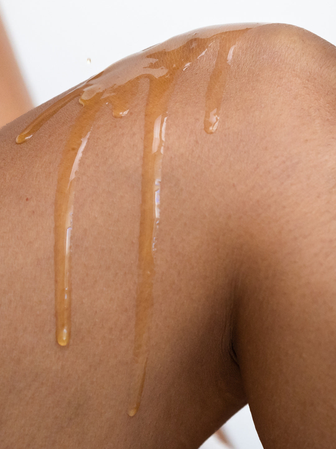 Close-up of golden Biologi Hydrate Body Oil running over a bare shoulder, showing the oil's sheen and fluid consistency on skin.