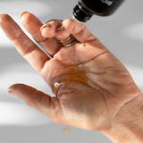 Close-up of Biologi Hydrate Body Oil being poured from its black bottle into an open palm, with a small stream of golden oil visible.