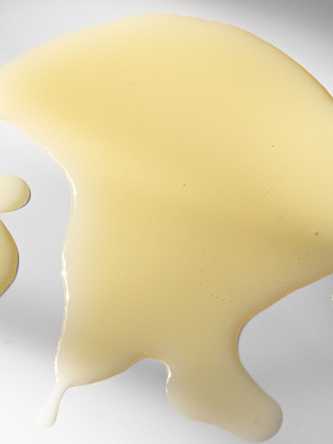 A broad pool of pale golden Biologi Hydrate Body Oil with a smaller round drop on white, showing its liquid texture and colour.