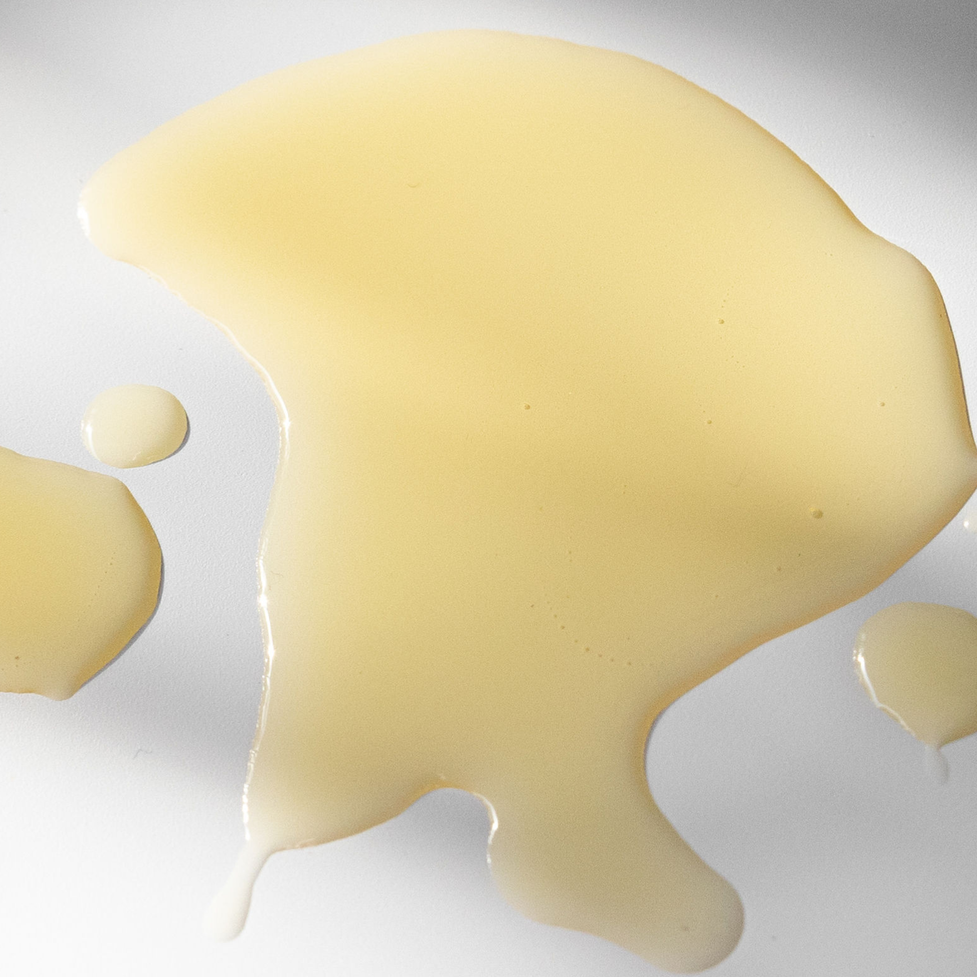 A broad pool of pale golden Biologi Hydrate Body Oil with a smaller round drop on white, showing its liquid texture and colour.