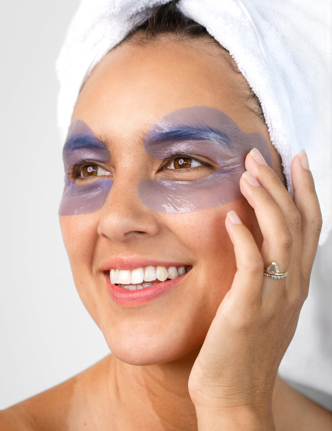 A woman with a white towel around her hair wears the lavender-blue Biologi Revive Bio Cellulose Eye Mask patches around both eyes, touching one patch gently.