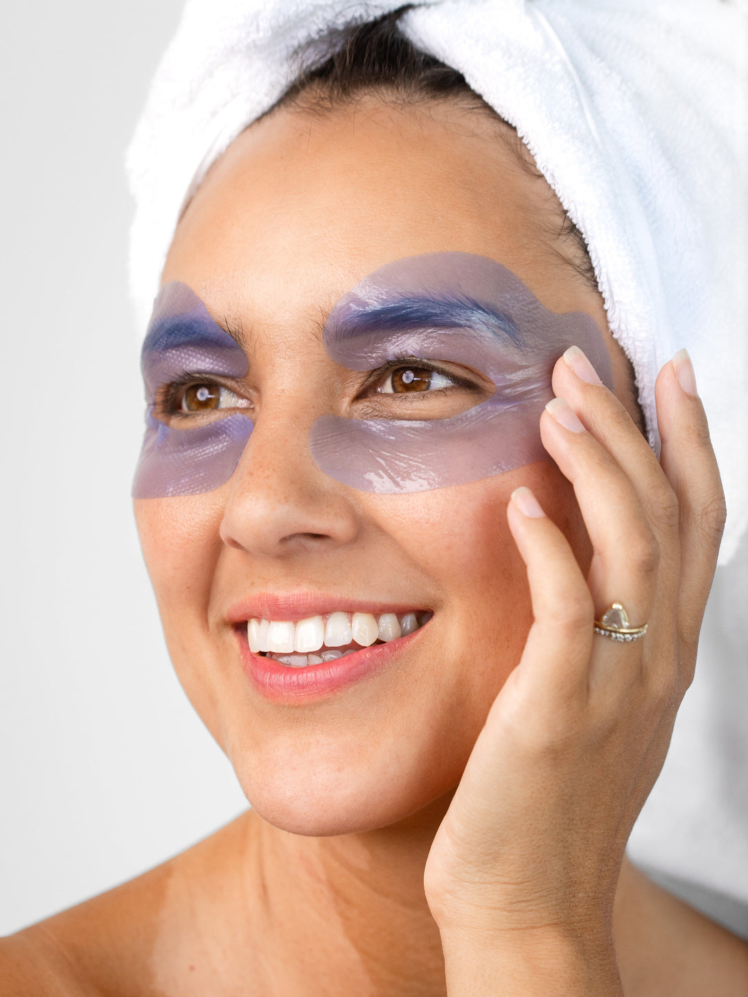 A woman with a white towel around her hair wears the lavender-blue Biologi Revive Bio Cellulose Eye Mask patches around both eyes, touching one patch gently.
