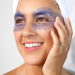 A woman with a white towel around her hair wears the lavender-blue Biologi Revive Bio Cellulose Eye Mask patches around both eyes, touching one patch gently.