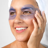 A woman with a white towel around her hair wears the lavender-blue Biologi Revive Bio Cellulose Eye Mask patches around both eyes, touching one patch gently.