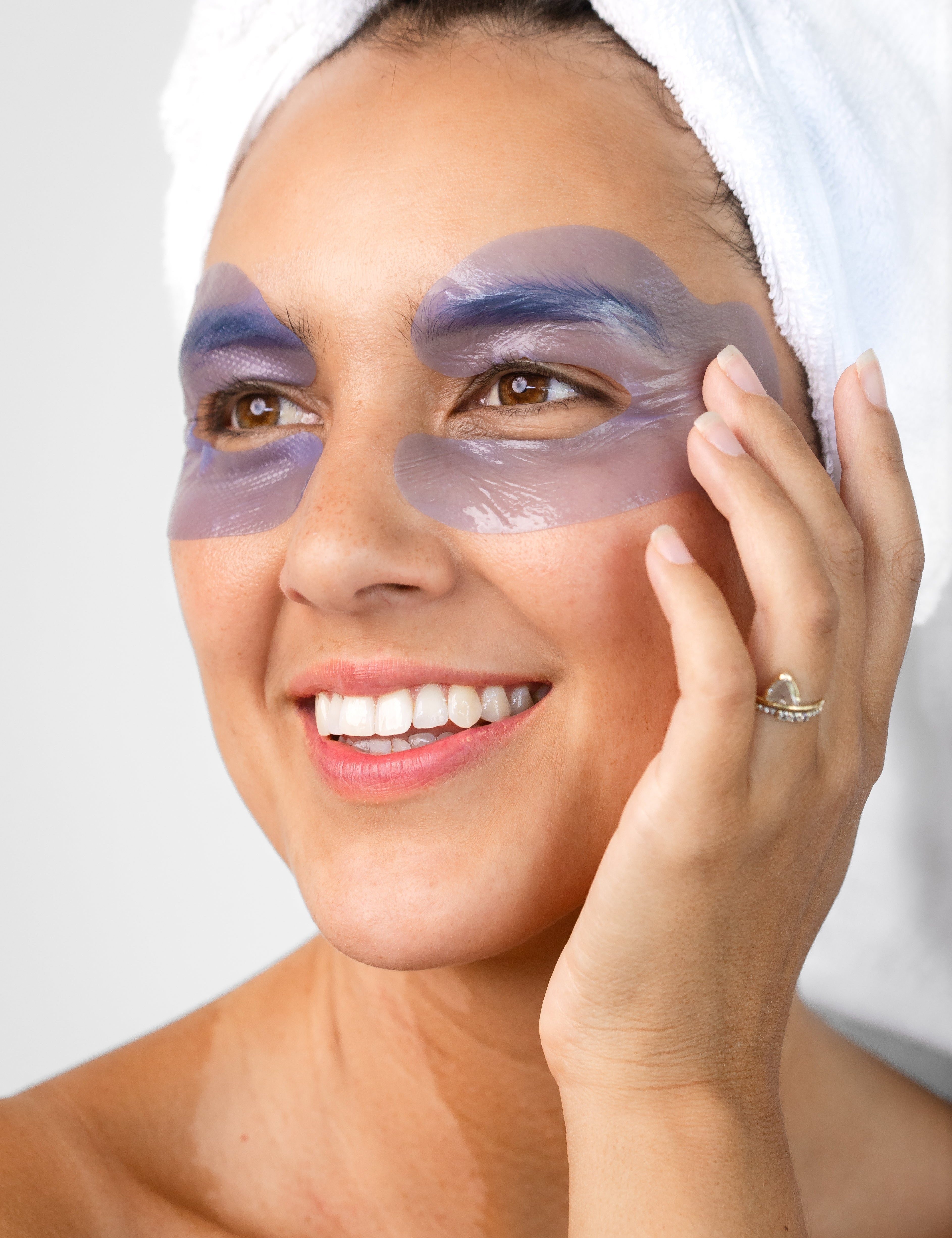 A woman with a white towel around her hair wears the lavender-blue Biologi Revive Bio Cellulose Eye Mask patches around both eyes, touching one patch gently.