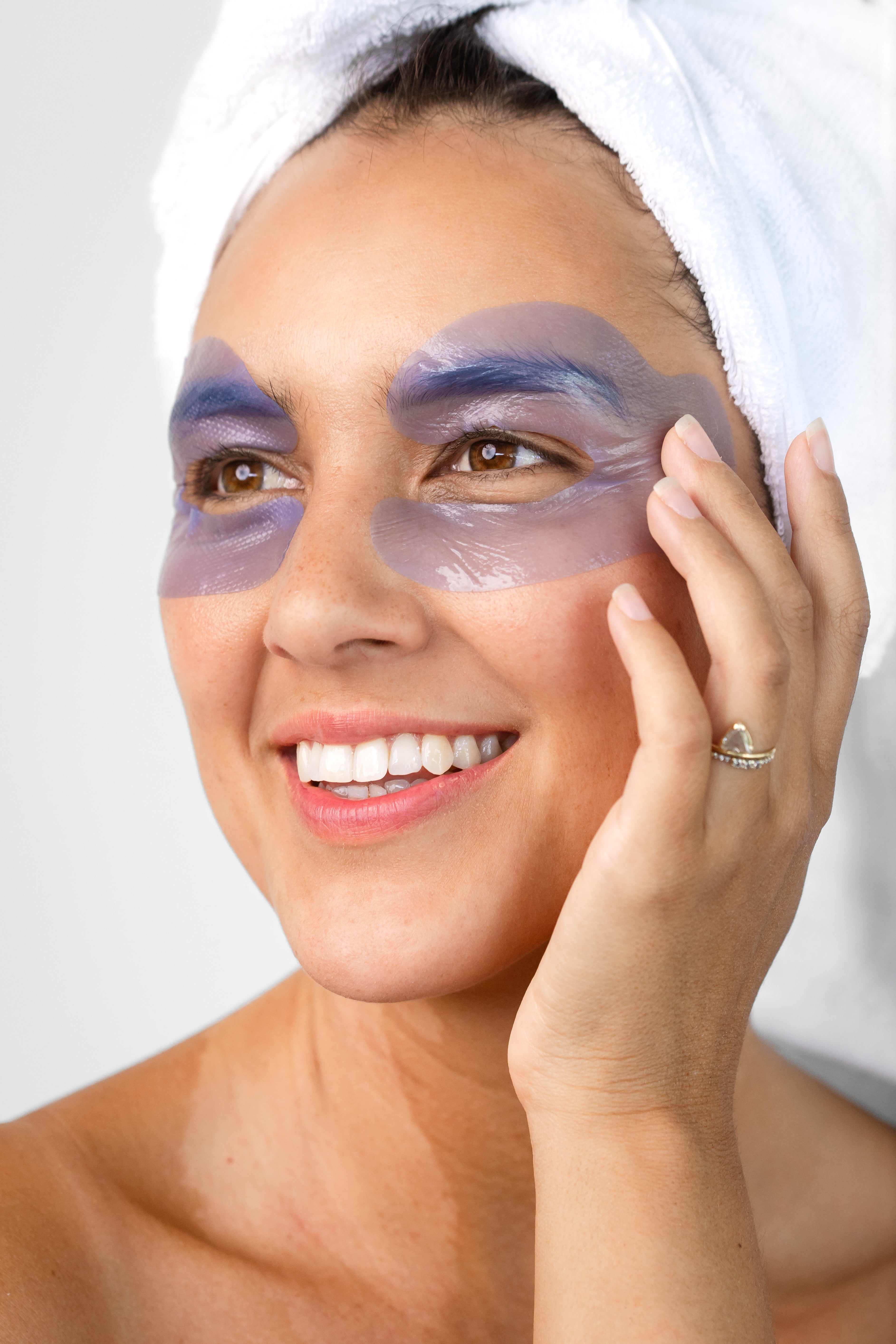 A woman with a white towel around her hair wears the lavender-blue Biologi Revive Bio Cellulose Eye Mask patches around both eyes, touching one patch gently.
