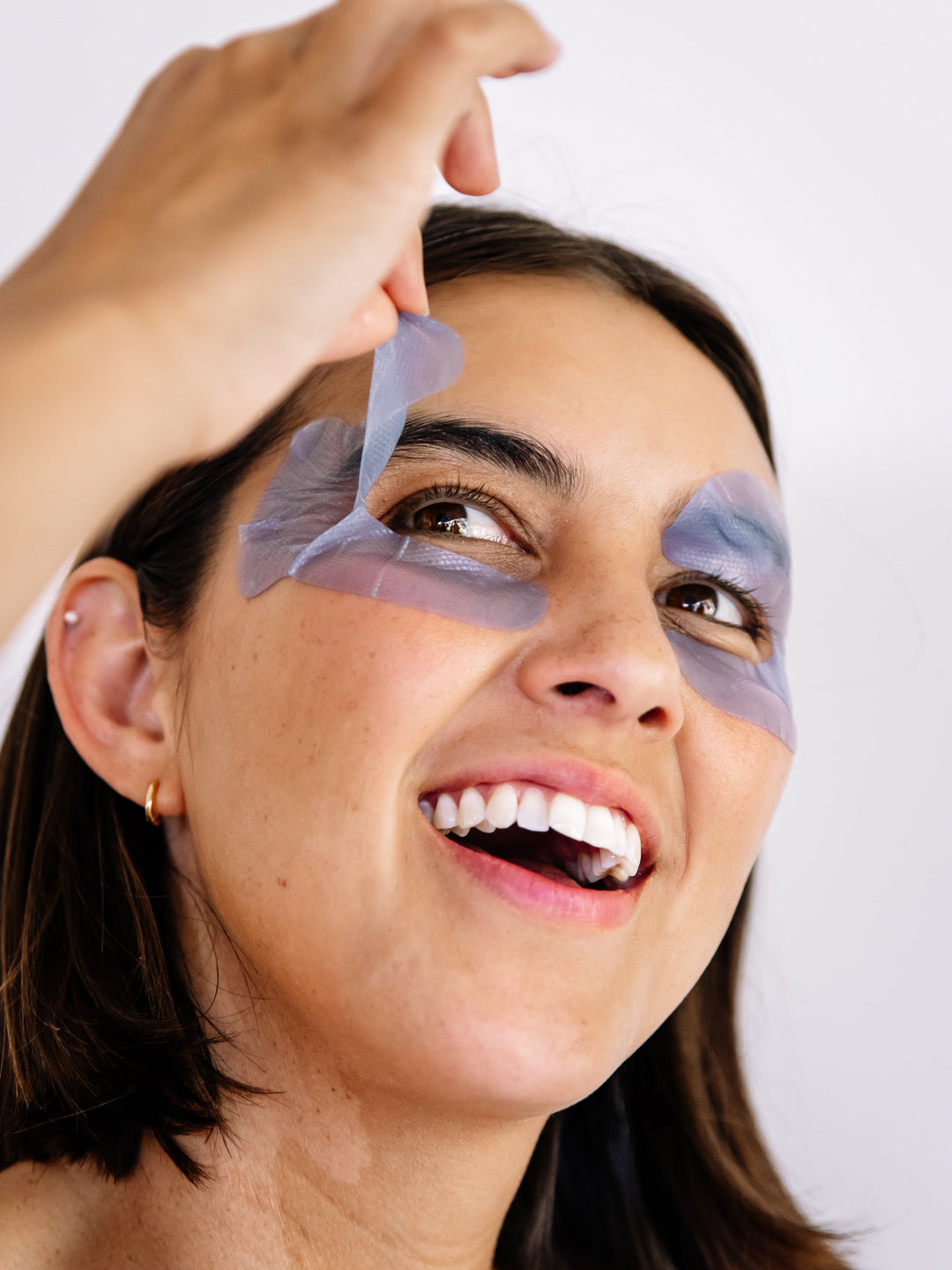 Close-up of a smiling woman lifting a lavender-blue Biologi Revive Bio Cellulose Eye Mask patch from around her eye.