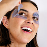 Close-up of a smiling woman lifting a lavender-blue Biologi Revive Bio Cellulose Eye Mask patch from around her eye.