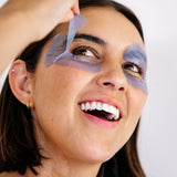 Close-up of a smiling woman lifting a lavender-blue Biologi Revive Bio Cellulose Eye Mask patch from around her eye.