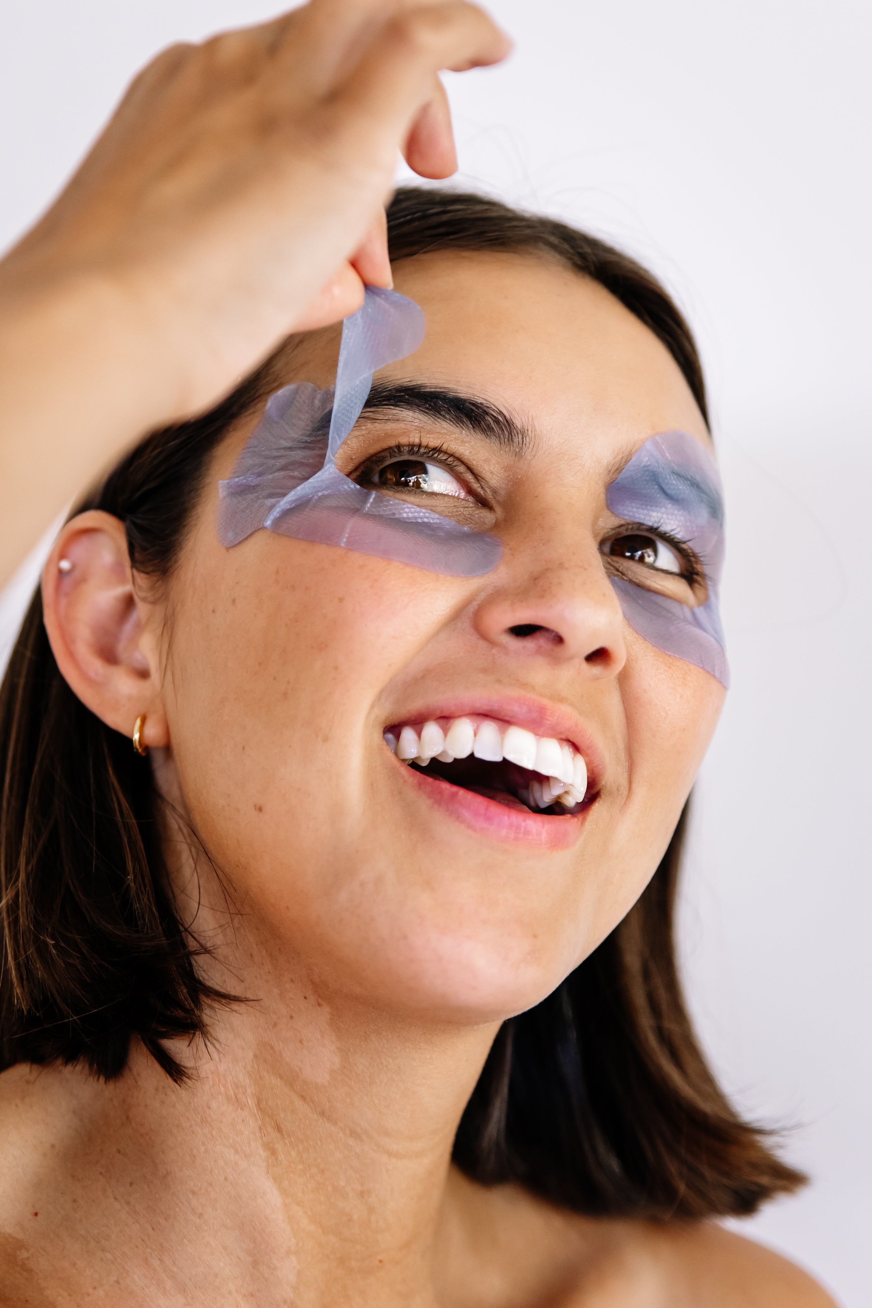 Close-up of a smiling woman lifting a lavender-blue Biologi Revive Bio Cellulose Eye Mask patch from around her eye.