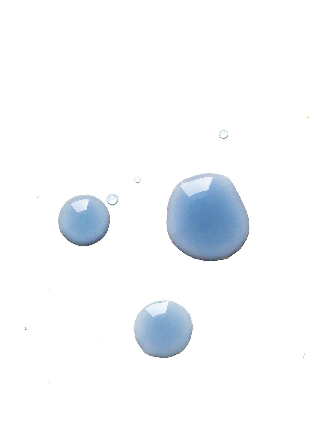 Three glossy blue-lavender drops of serum on white, illustrating the colour and fluid texture of Biologi Revive Bio Cellulose Eye Mask essence.
