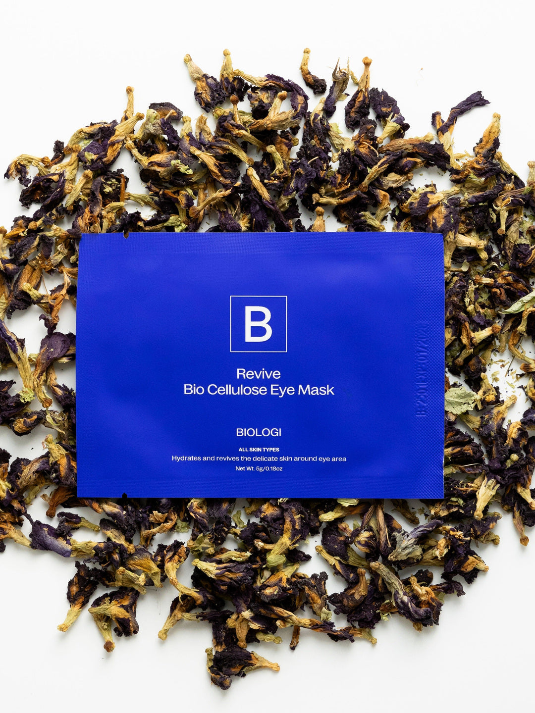 A cobalt-blue Biologi Revive Bio Cellulose Eye Mask sachet surrounded by dried butterfly pea flowers on white.