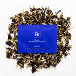 A cobalt-blue Biologi Revive Bio Cellulose Eye Mask sachet surrounded by dried butterfly pea flowers on white.