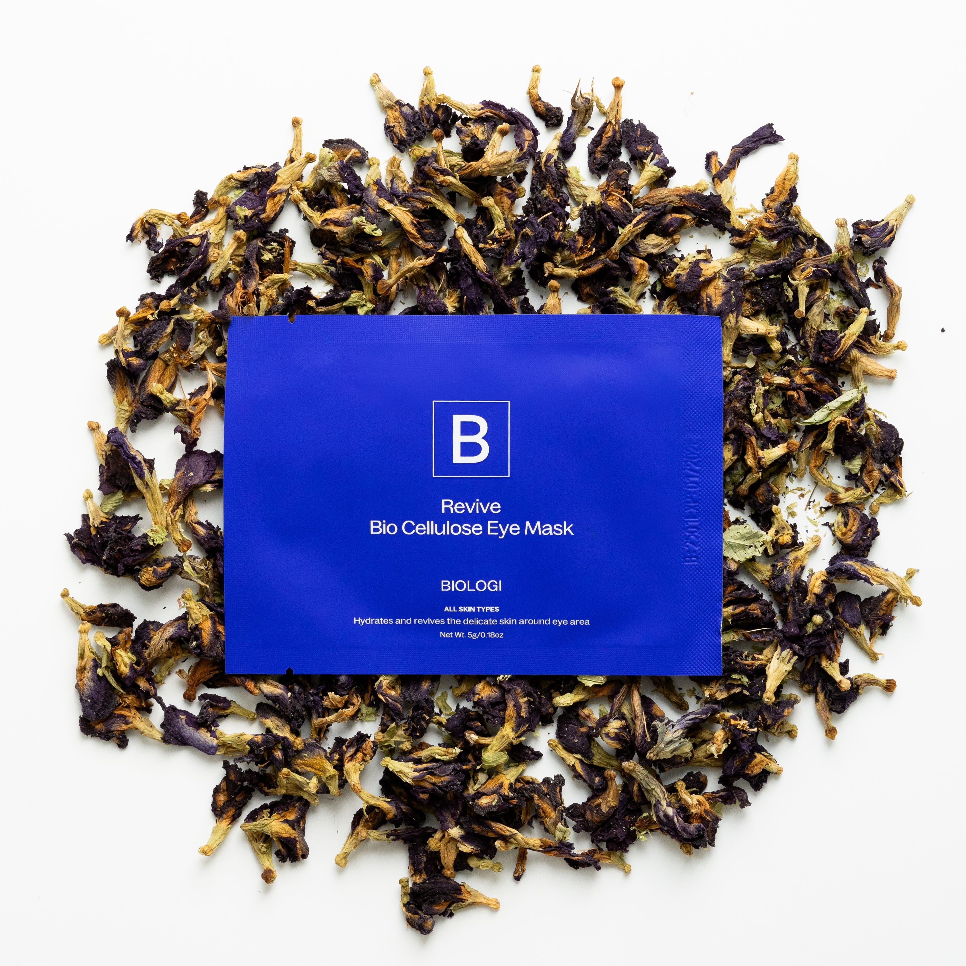 A cobalt-blue Biologi Revive Bio Cellulose Eye Mask sachet surrounded by dried butterfly pea flowers on white.