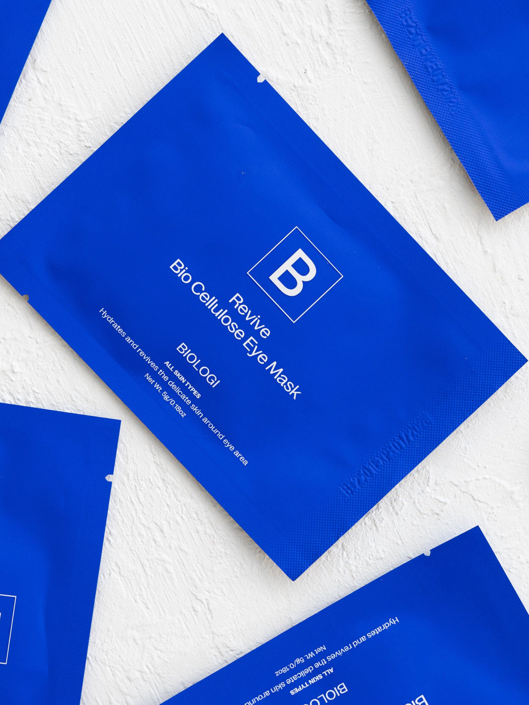 Several cobalt-blue Biologi Revive Bio Cellulose Eye Mask sachets arranged diagonally on a textured white fabric surface.