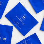Several cobalt-blue Biologi Revive Bio Cellulose Eye Mask sachets arranged diagonally on a textured white fabric surface.