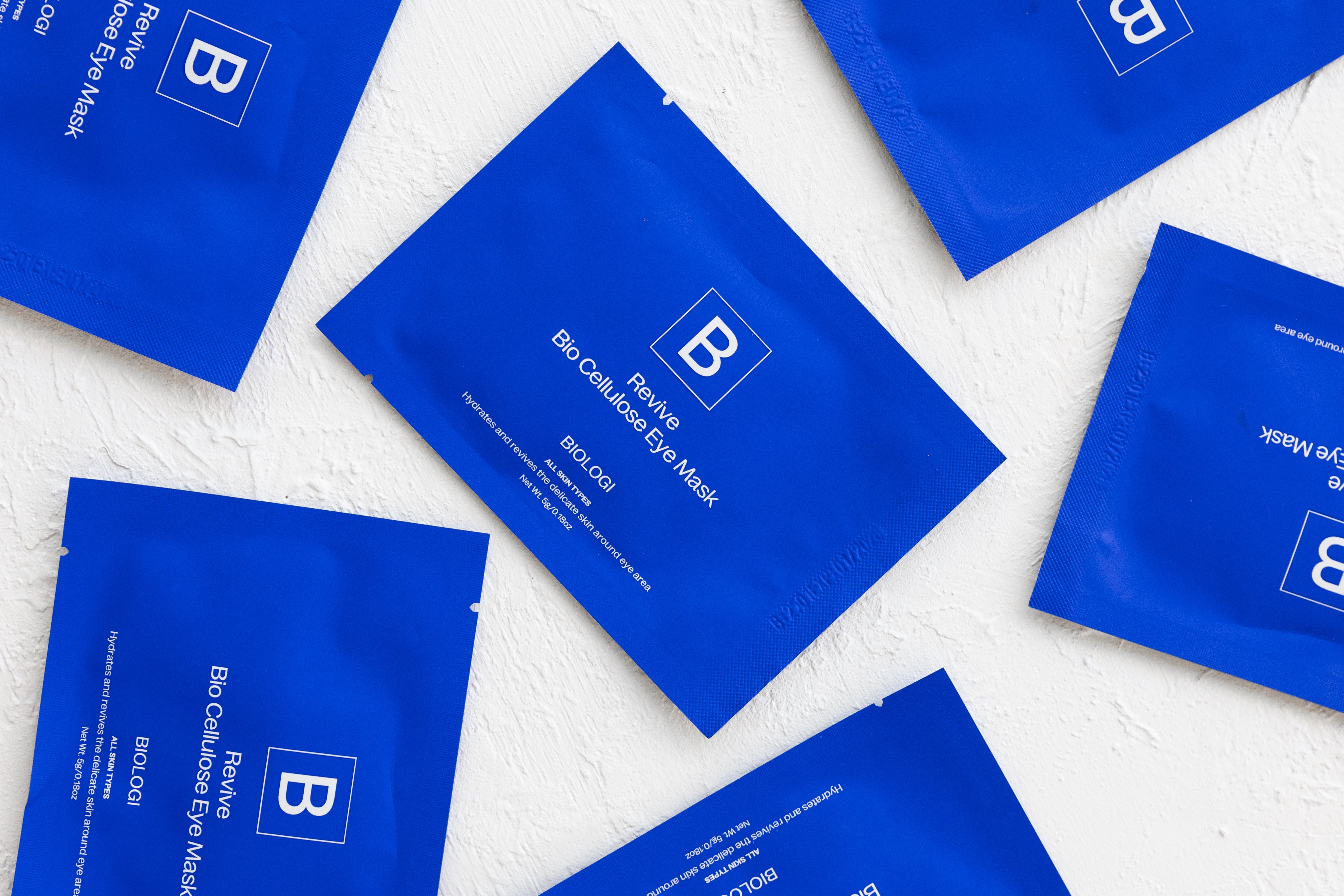 Several cobalt-blue Biologi Revive Bio Cellulose Eye Mask sachets arranged diagonally on a textured white fabric surface.
