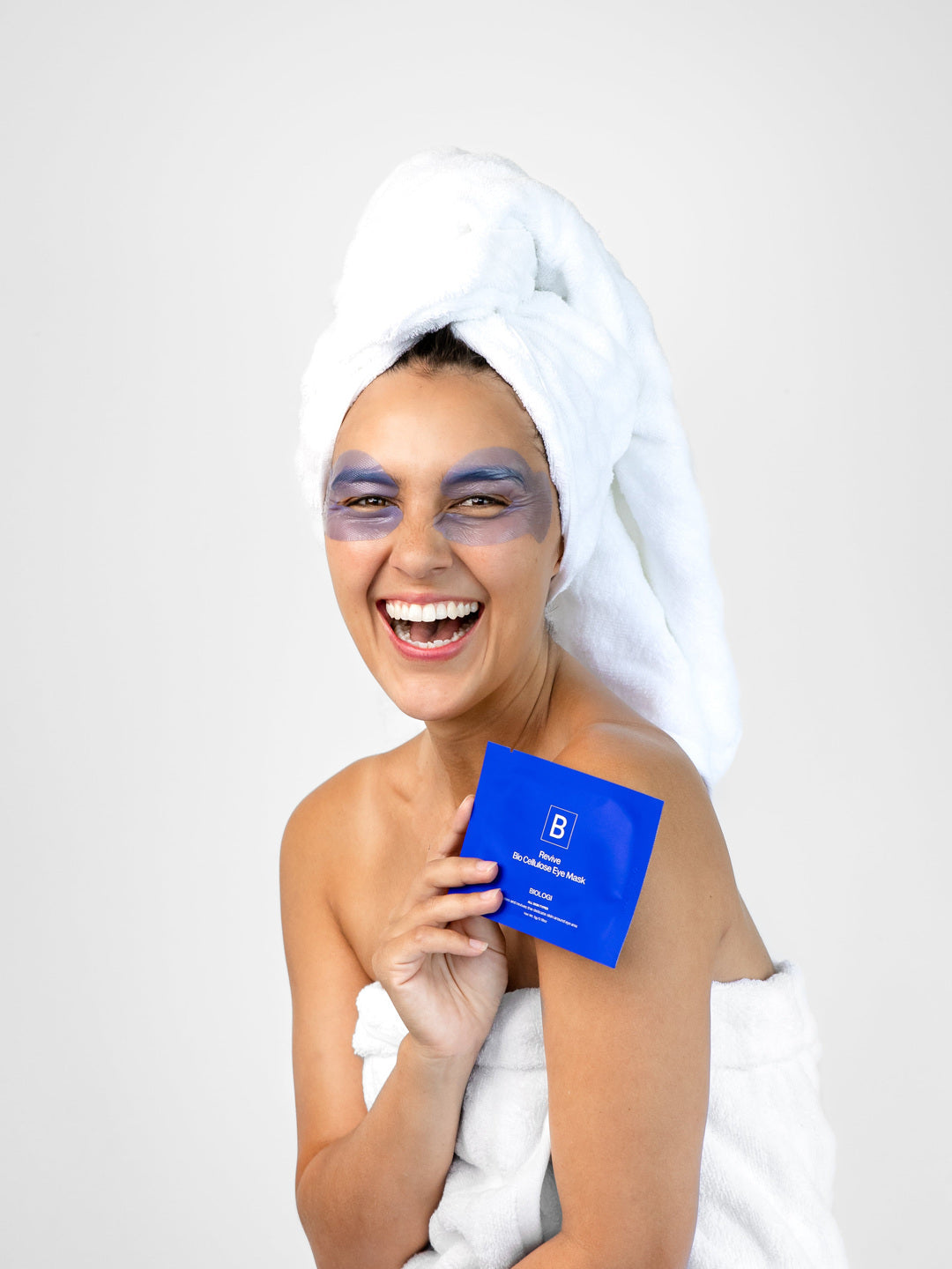 A smiling woman in a white towel wears lavender-blue eye mask patches and holds a cobalt-blue Biologi Revive Bio Cellulose Eye Mask sachet.
