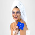 A smiling woman in a white towel wears lavender-blue eye mask patches and holds a cobalt-blue Biologi Revive Bio Cellulose Eye Mask sachet.