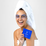 A smiling woman in a white towel wears lavender-blue eye mask patches and holds a cobalt-blue Biologi Revive Bio Cellulose Eye Mask sachet.