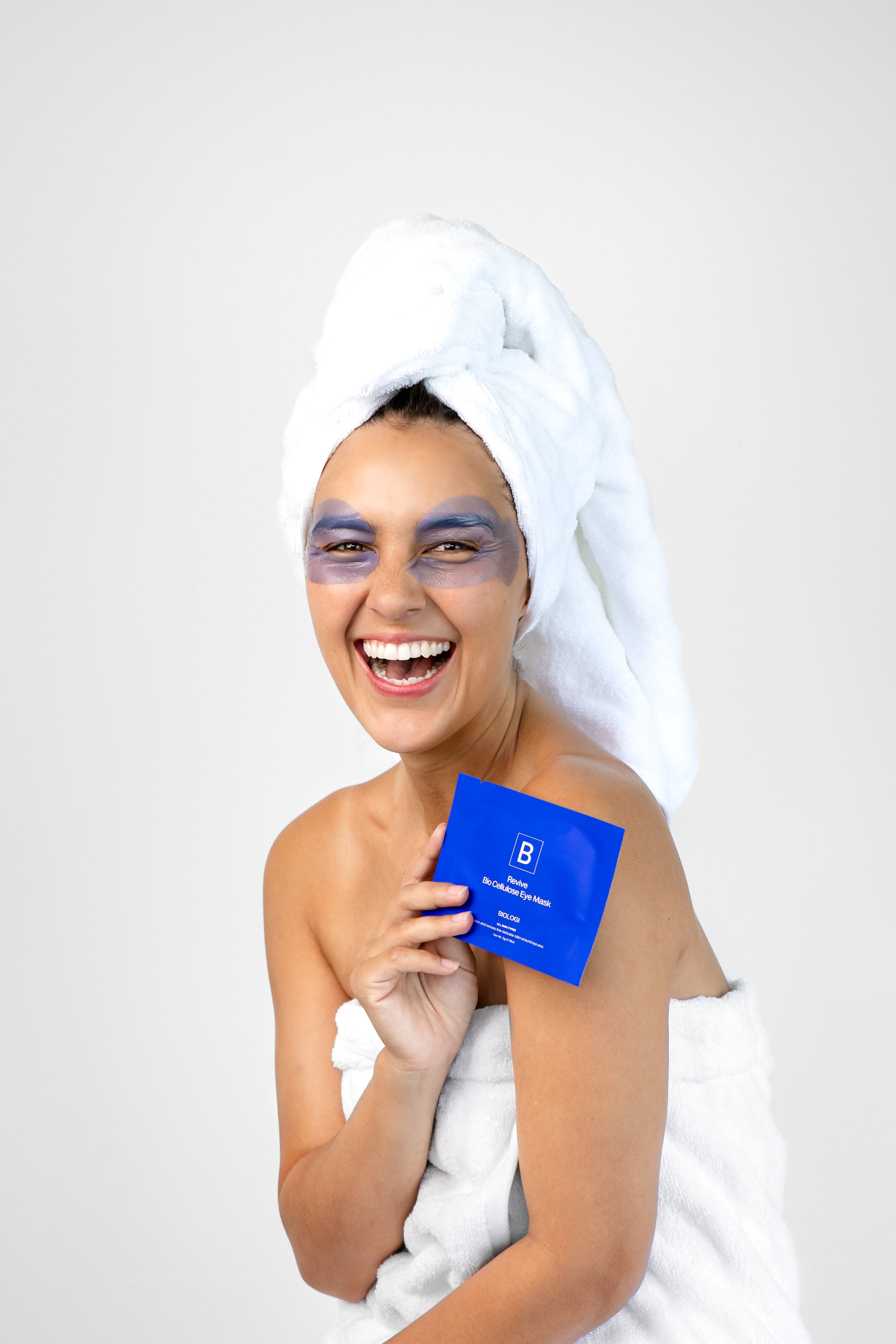 A smiling woman in a white towel wears lavender-blue eye mask patches and holds a cobalt-blue Biologi Revive Bio Cellulose Eye Mask sachet.