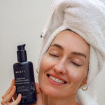 Woman in a towel turban smiling with eyes closed holding Mukti Hydrating Cleansing Lotion